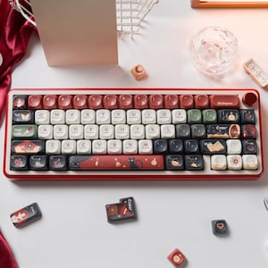 May include: A mechanical keyboard with a red frame and a multi-colored keycap set featuring various illustrations including a black cat, potions, and stars. The keys include standard letters, numbers, and symbols, with specific keys labeled Backspace and Enter.