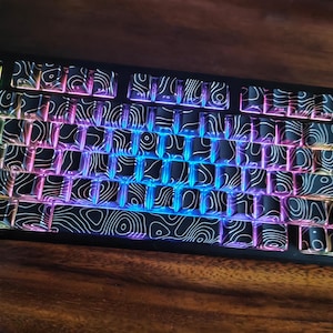 May include: A black mechanical keyboard with keycaps featuring a white topographic pattern. The keys are illuminated with a rainbow gradient of colors, including blue, purple, and green. The keyboard sits on a dark wood surface.