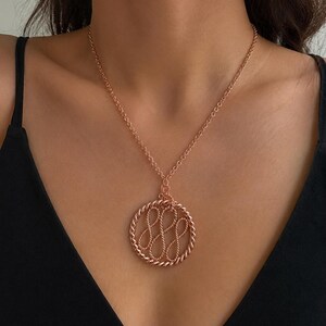 May include: Rose gold necklace featuring a circular pendant with a twisted rope border and an inner design of intertwined loops. The necklace is worn against a black top, highlighting the pendant's intricate details and warm color.