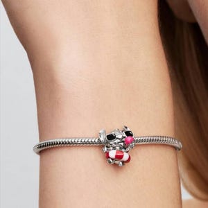 May include: A silver snake chain bracelet featuring a charm of a character with black sunglasses and a pink nose, holding a red and white striped life preserver. The charm is crafted from polished silver metal with colorful enamel detailing.