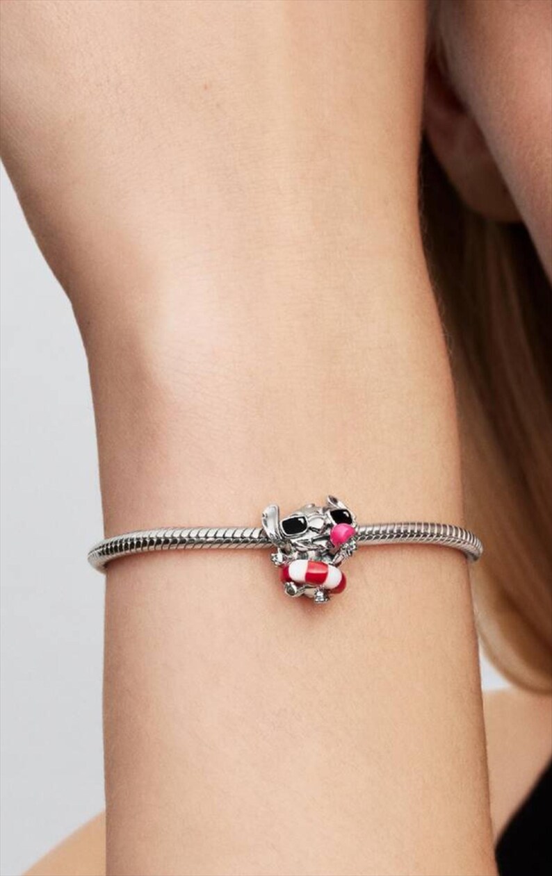 May include: A silver snake chain bracelet featuring a charm of a character with black sunglasses and a pink nose, holding a red and white striped life preserver. The charm is crafted from polished silver metal with colorful enamel detailing.