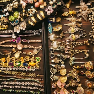 May include: A collection of assorted jewellery including gold-tone bracelets, necklaces, and earrings arranged in black display trays. The items feature various colourful gemstones, floral motifs, metallic finishes, and intricate link designs.