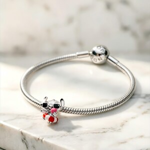 May include: A silver snake chain bracelet featuring a charm of a character wearing black sunglasses and a red and white striped life preserver, while holding a small pink object. The bracelet has a round clasp with the engraved text PANDORA.