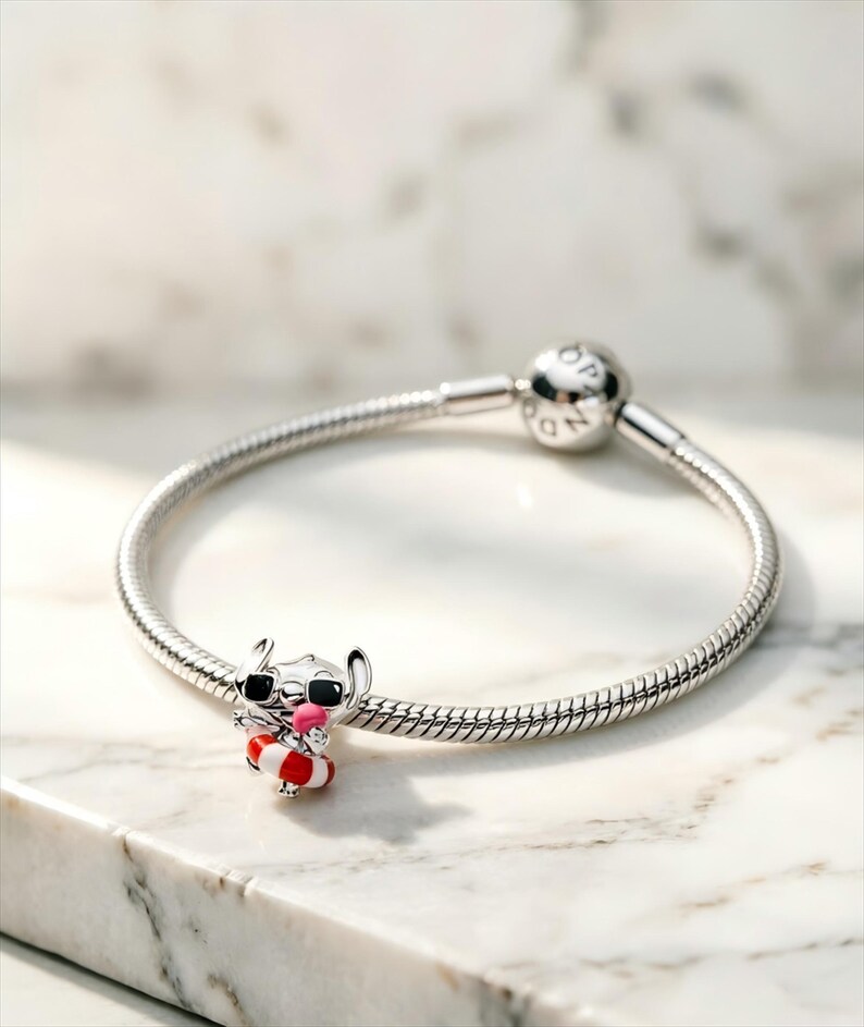 May include: A silver snake chain bracelet featuring a charm of a character wearing black sunglasses and a red and white striped life preserver, while holding a small pink object. The bracelet has a round clasp with the engraved text PANDORA.