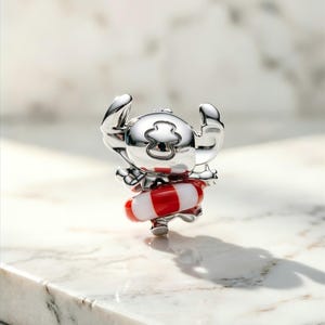 May include: A polished silver-toned charm featuring a character with large ears and a head marking, wearing a red and white striped life preserver ring around its waist. The metallic surface reflects light, highlighting the smooth, rounded contours.