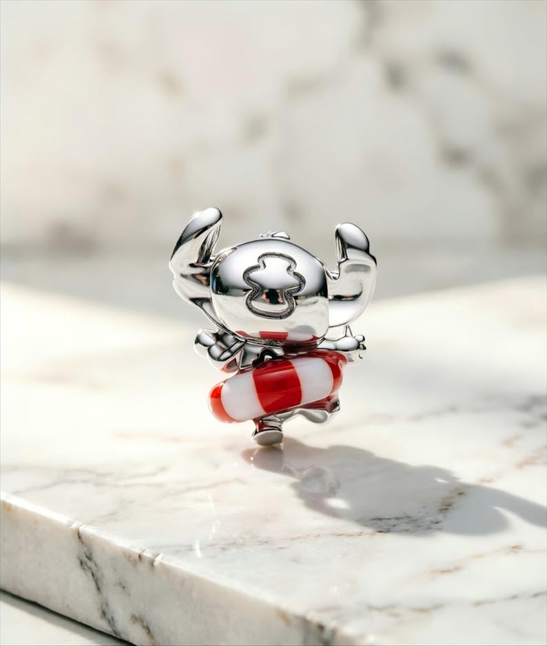 May include: A polished silver-toned charm featuring a character with large ears and a head marking, wearing a red and white striped life preserver ring around its waist. The metallic surface reflects light, highlighting the smooth, rounded contours.