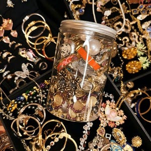 May include: A clear jar filled with various jewelry pieces, including necklaces, bracelets, and earrings. The jewelry is a mix of gold, silver, and colorful accents, with pearls and other decorative elements. The pieces are arranged on a black surface.