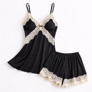 May include: Black satin pajama set with a camisole top and shorts. The camisole features a V-neckline with cream-colored lace trim and a small bow. The shorts have matching lace trim along the hem.