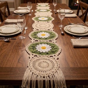 May include: A long, cream-colored crochet table runner featuring alternating circular patterns and oval sections with white and yellow daisy motifs on a green background. The runner ends with long, dangling fringe tassels at both ends of the piece.