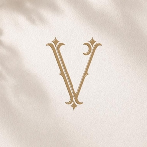 Può includere: Un monogramma color oro della lettera "V" su una carta color crema. La parola "MONOGRAM" è in alto e "Monograms Logos Alwih" in basso. Una penna d'oro, un nastro e dei fiori sono sullo sfondo.