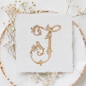 Könnte beinhalten: Ein goldfarbenes Monogramm des Buchstabens "J" mit kunstvollen floralen Details, dargestellt auf strukturiertem weißem Papier. Das Papier liegt auf einem weißen Teller mit Goldrand, umgeben von zarten weißen Blumen. Das Wort "MONOGRAMM" steht oben.