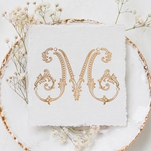 Puede incluir: Un monograma dorado con la letra "M" y adornos ornamentales, exhibido sobre papel blanco texturizado. El papel se encuentra sobre un plato blanco con borde dorado, adornado con delicadas flores blancas. La palabra "MONOGRAMA" está en la parte superior.