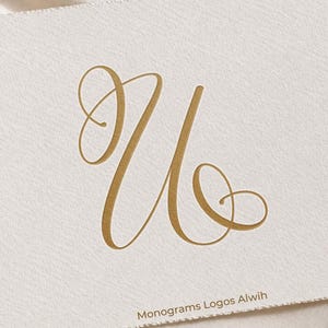 Könnte beinhalten: Eine cremefarbene Karte zeigt den eleganten, goldfarbenen Buchstaben "U" in einer fließenden Schrift. Die Karte hat eine strukturierte Oberfläche und der Text "Monograms Logos Alwih" ist unten aufgedruckt.