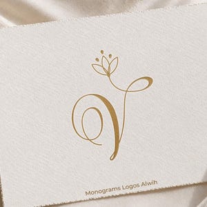 Può includere: Un monogramma color oro con un elemento floreale stilizzato sopra una lettera "V" fluida. Stampato su una carta strutturata bianco sporco. Il testo "Monograms Logos Alwih" è stampato in basso.