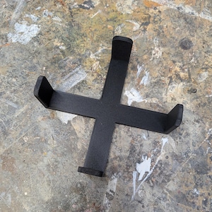 May include: A black, cross-shaped object with four arms, each ending in a raised, rectangular support. The object appears to be 3D-printed and sits on a mottled surface with shades of grey, brown, and white.