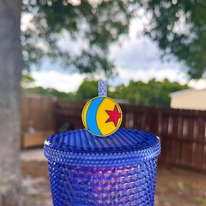 May include: A blue tumbler with a textured surface, topped with a charm featuring a yellow ball with a blue stripe and a red star. The charm is attached to a straw.