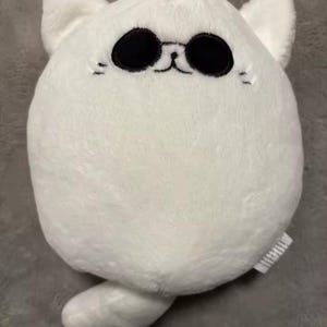 May include: A white plush cat toy with black oval eyes, small black whiskers, and a stitched mouth. The cat has pointed ears and a small tail. The toy is made of soft, fluffy material.