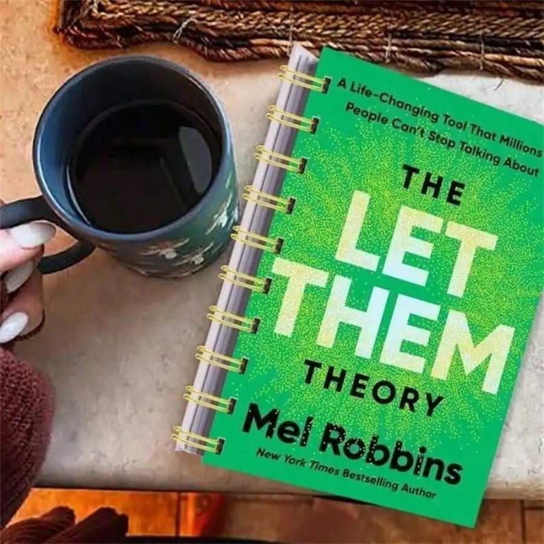May include: A hardcover book titled "THE LET THEM THEORY" by Mel Robbins, with a bright green cover and gold spiral binding. A dark blue mug with a floral design sits next to the book, filled with a dark liquid.