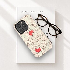 Op de afbeelding: Een telefoonhoesje met een crèmekleurige achtergrond en een patroon van rode en gestippelde harten. De hoes ligt op een wit boek met de tekst "Stuffed with listings and tales". Een bril met schildpadmotief ligt op het boek.