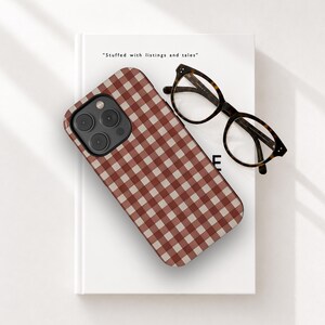 May include: A phone case with a red and beige gingham pattern rests on a white book. The phone case has a black camera module. Tortoiseshell eyeglasses are next to the phone case. The book has the text "Stuffed with listings and tales".