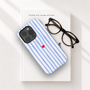 Peut inclure: Coque de téléphone à rayures bleues et blanches avec un petit cœur rouge. La coque est posée sur un livre blanc avec le texte "Stuffed with listings and tales". Des lunettes en écaille sont à côté.