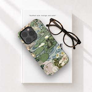 Puede incluir: Una funda de teléfono con un diseño temático de la naturaleza que presenta una rana en un nenúfar, flores blancas y una escena acuática. La funda está sobre un libro blanco con el texto "Stuffed with listings and tales". Gafas carey descansan sobre el libro.