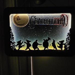 May include: A rectangular, illuminated wall art piece featuring a night sky scene. The design includes a crescent moon, the words "Grateful Dead", and dancing bear silhouettes. The frame is black.