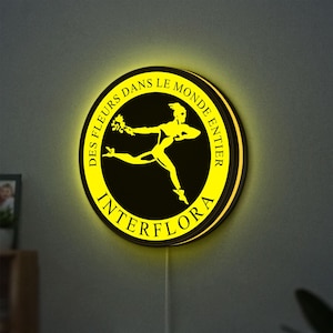 May include: A circular illuminated sign with a black centre and yellow text and a running figure. The text reads "DES FLEURS DANS LE MONDE ENTIER" and "INTERFLORA". The sign is mounted on a grey wall.