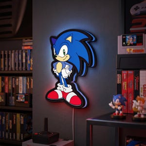 Op de afbeelding: Een blauwe, zwarte en rode Sonic the Hedgehog wandlamp. Het licht heeft de vorm van het personage en heeft een gloeiende omtrek. Het licht is aangesloten op een stopcontact met een wit snoer. De achtergrond bevat gameconsoles en game cartridges.