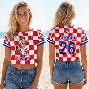 May include: A short-sleeved crop top featuring a red and white checkered pattern. The front displays a cartoon animal wearing a matching jersey with a soccer ball and the word Custom. The back shows the word Custom and the number 26 in blue.