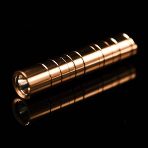 May include: A cylindrical copper-colored flashlight with a smooth, polished metallic finish. The body features a series of evenly spaced, recessed horizontal grooves along its length, and the front end shows a circular opening with a reflective lens.