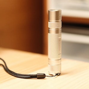 May include: A silver flashlight with a textured grip and a black lanyard. The flashlight is cylindrical with a ribbed design on the body. It is resting on a light-coloured wooden surface.