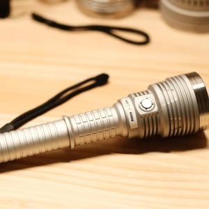 Puede incluir: Una linterna plateada con un cordón negro descansa sobre una superficie de madera. La linterna tiene un mango texturizado y una gran cabeza circular. El texto "ADJ 317 6000K" está impreso en el lateral. La linterna está diseñada para la iluminación.