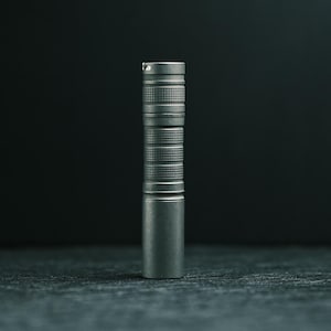 May include: A cylindrical metal flashlight with a matte gray finish. The body features two distinct sections with a fine, textured cross-hatch knurling pattern for grip, while the base and top sections remain smooth and unadorned in design.
