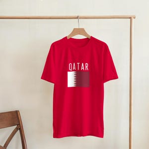 May include: A red t-shirt displaying the word "QATAR" in white above a graphic of the Qatar flag. The flag design has a white serrated edge on the left and a maroon rectangle on the right. The shirt is suspended from a wooden clothes rail.