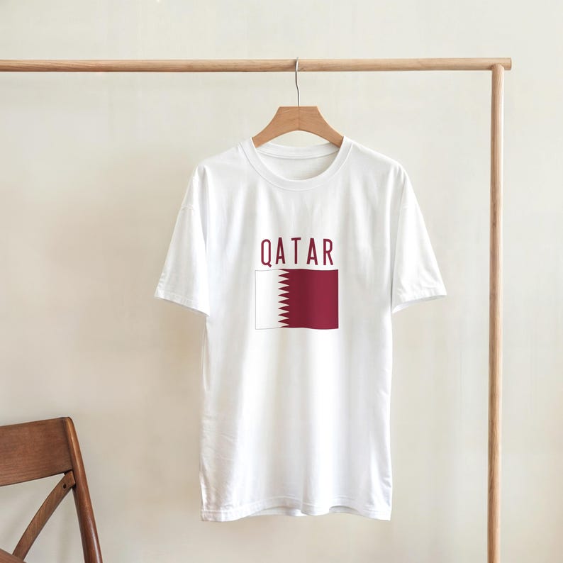 May include: White t-shirt featuring the word "QATAR" in maroon above a maroon and white flag design. The t-shirt is displayed on a wooden hanger against a plain backdrop.