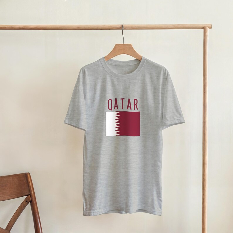 May include: A grey t-shirt featuring the word "QATAR" in maroon above a maroon and white flag design. The shirt is suspended on a wooden hanger from a metal clothing rail.