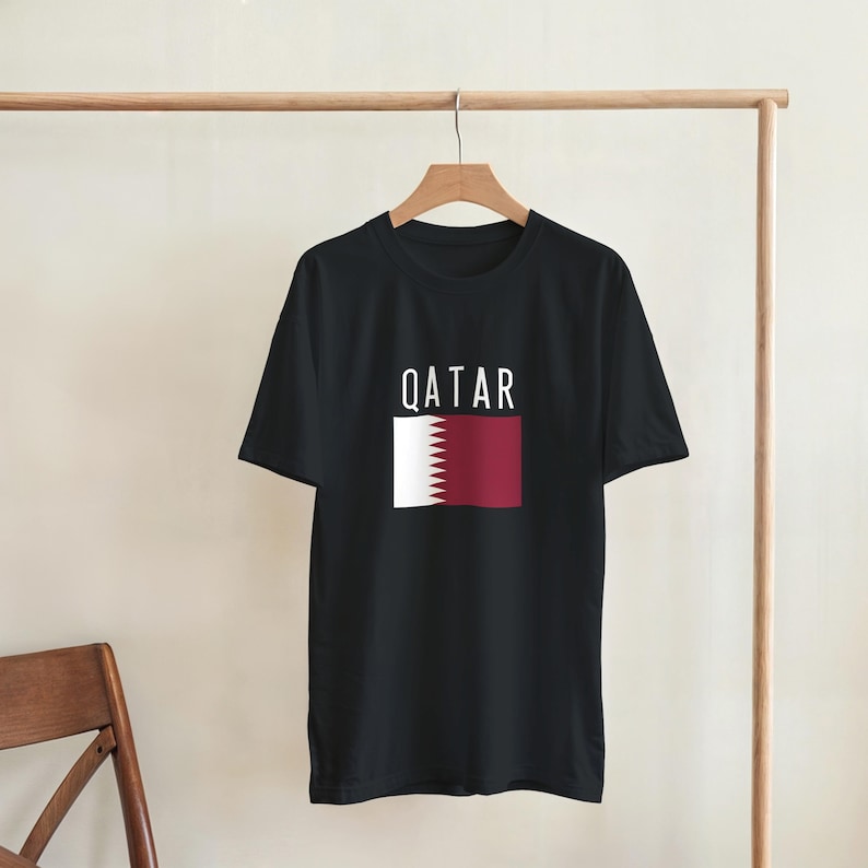 May include: Black t-shirt displaying the word "QATAR" in white capital letters above a graphic of the Qatar flag. The flag design features a white serrated edge and a maroon rectangle.