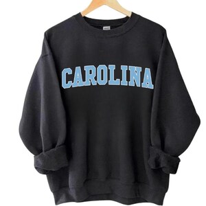 May include: Black sweatshirt featuring the word "CAROLINA" in light blue block lettering. The garment has a crew neck and long sleeves, with the cuffs rolled up. The sweatshirt is hanging on a wooden hanger.