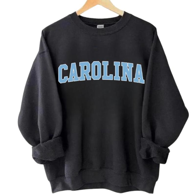May include: Black sweatshirt featuring the word "CAROLINA" in light blue block lettering. The garment has a crew neck and long sleeves, with the cuffs rolled up. The sweatshirt is hanging on a wooden hanger.