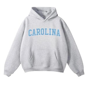 May include: A light grey hooded sweatshirt featuring the word "CAROLINA" in light blue block lettering across the chest. The hoodie has a hood and a front pocket. The material looks soft and comfortable.