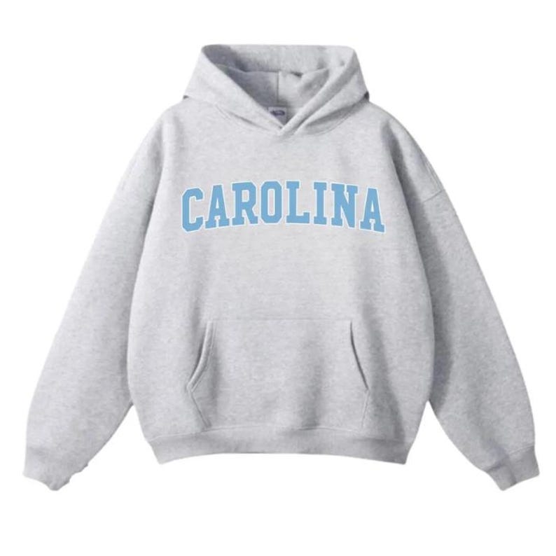 May include: A light grey hooded sweatshirt featuring the word "CAROLINA" in light blue block lettering across the chest. The hoodie has a hood and a front pocket. The material looks soft and comfortable.