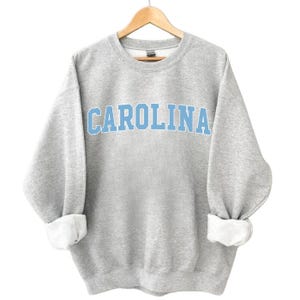 May include: A grey sweatshirt with the word "CAROLINA" in light blue block letters across the chest. The sweatshirt has a crew neck, long sleeves, and folded cuffs. The garment is hanging on a wooden hanger.