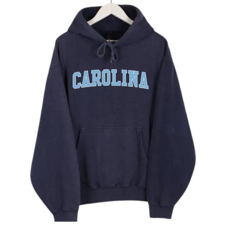 May include: A navy blue hooded sweatshirt with the word "CAROLINA" in light blue block letters across the chest. The sweatshirt has a front pocket and a drawstring hood. The garment is hanging on a white hanger.