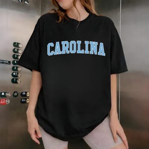 May include: Black t-shirt with the word "CAROLINA" in light blue block letters across the chest. The shirt has short sleeves and a crew neck. The model is wearing light-coloured shorts.