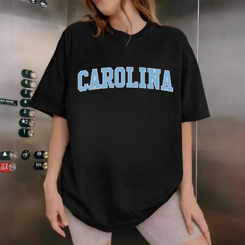 May include: Black t-shirt with the word "CAROLINA" in light blue block letters across the chest. The shirt has short sleeves and a crew neck. The model is wearing light-coloured shorts.