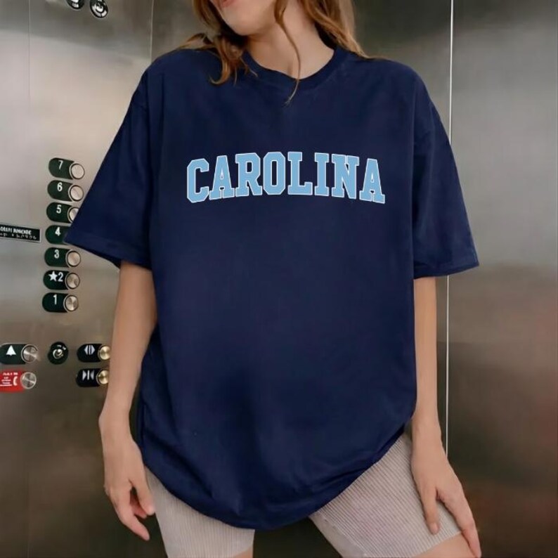 May include: A navy blue t-shirt featuring the word "CAROLINA" in light blue block lettering. The top has a crew neck and short sleeves. The model is wearing beige shorts.