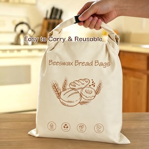 Op de afbeelding: Beige bijenwas broodzak met een handvat en zwarte gesp. De tas heeft de tekst "Easy to Carry & Reusable" en "Beeswax Bread Bags". Een bruine illustratie van brood en tarwe is op de tas gedrukt.