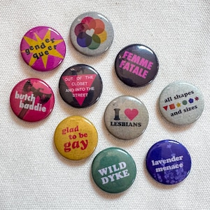 May include: A collection of nine circular pin-back buttons featuring various designs and text, including phrases like gender queer, femme fatale, butch baddie, out of the closet and into the street, I love lesbians, all shapes and sizes, glad to be gay, wild dyke, and lavender menace.