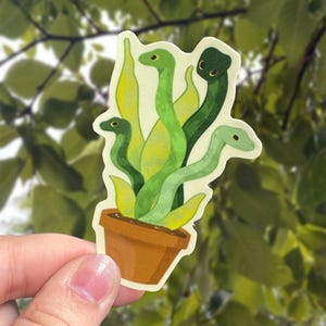 May include: A die-cut sticker featuring a brown terracotta pot containing several green snakes that resemble the leaves of a snake plant. The snakes are depicted in various shades of green, ranging from light lime to dark forest green, with small eyes.